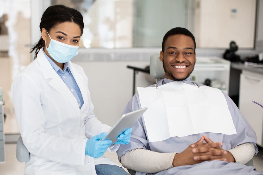 Dental Insurance Concept. Black Dentist Woman And Male Patient P A young man sitting in a dental chair with a dental assistant next to him.